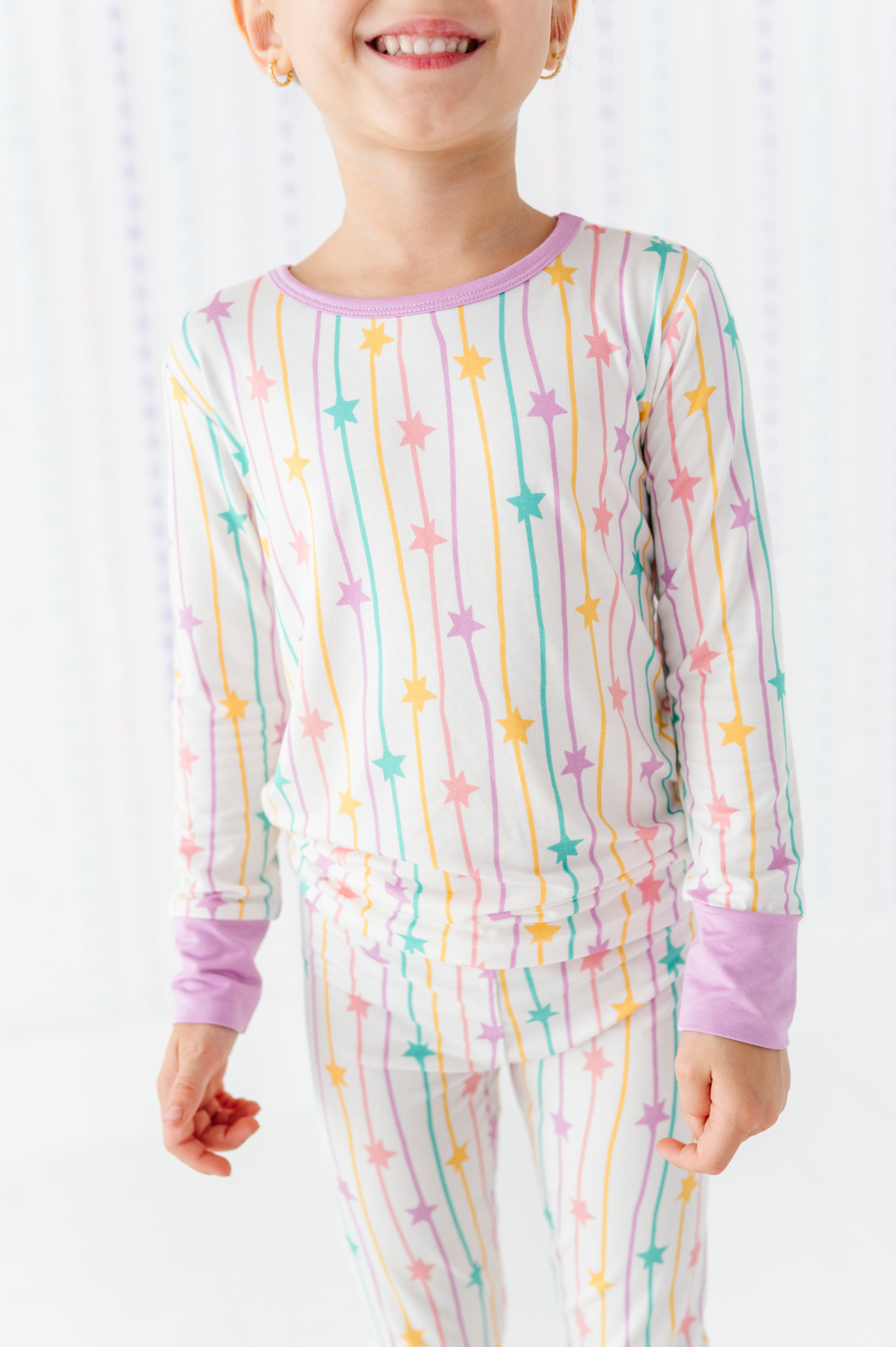 A child wearing a long-sleeve pajama top and matching pants with a pastel rainbow star-and-stripe print on a white background.